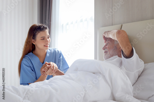 Smiling beautiful caring female doctor holding hand of male senior patient who lying in hospital bed. Nurse takes care elderly man feeling sick need to rest in bed at home, medical elderly health care © Stella
