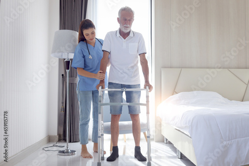 Female nurse or doctor helping elderly patient man lean to walk with orthopedic walker, patient practice walking inside house with physiotherapist, nursing senior people at house, medical health care. © Stella