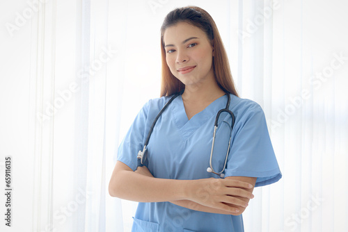 Portrait of happy smiling beautiful woman doctor with stethoscope in blue coat standing with arms crossed by window in clinic hospital. Female nurse on white background, medical health care worker. © Stella