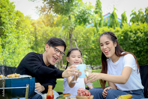 Joyful parent and kid child make barbecue at picnic party, have fun together in summer green garden, father mother and daughter girl enjoy eating and drinking at backyard, happy family love bonding.
