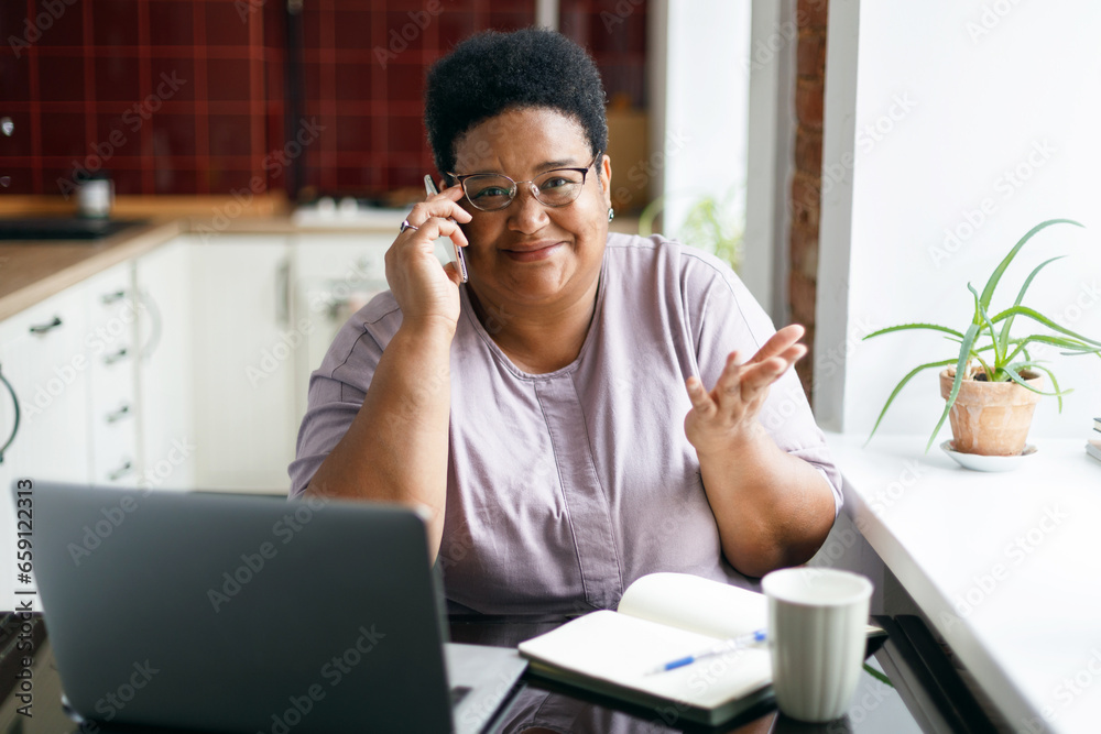 Puzzled senior overweight black female of talking on smartphone with phone scammers, looking at camera with confusion gesticulating shrugging her shoulders sitting at kitchen table in front of laptop Puzzled senior overweight black female of talking on smartphone with phone scammers, looking at camera with confusion gesticulating shrugging her shoulders sitting at kitchen table in front of laptop