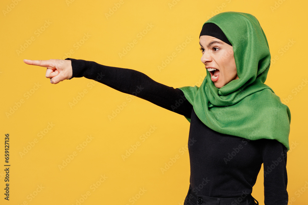 Side view young arabian asian muslim woman wear green hijab abaya black clothes point index finger aside scream isolated on plain yellow background. People uae middle eastern islam religious concept. Side view young arabian asian muslim woman wear green hijab abaya black clothes point index finger aside scream isolated on plain yellow background. People uae middle eastern islam religious concept.