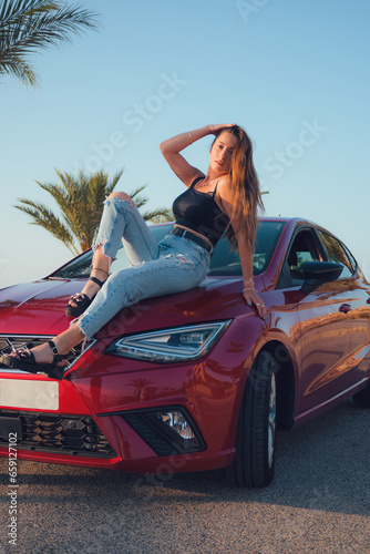 Beautiful woman posing above a red car at sunset