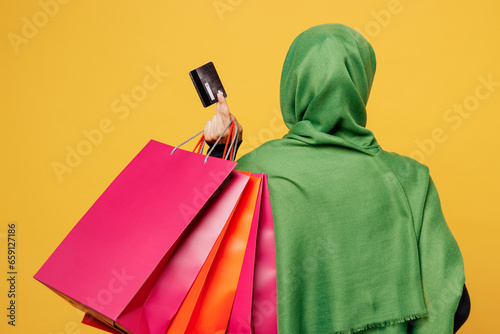 Back view young arab asian muslim woman wears green hijab abaya clothes hold shopping package bags credit bank card isolated on plain yellow background. Black Friday sale buy day, uae islam concept. photo