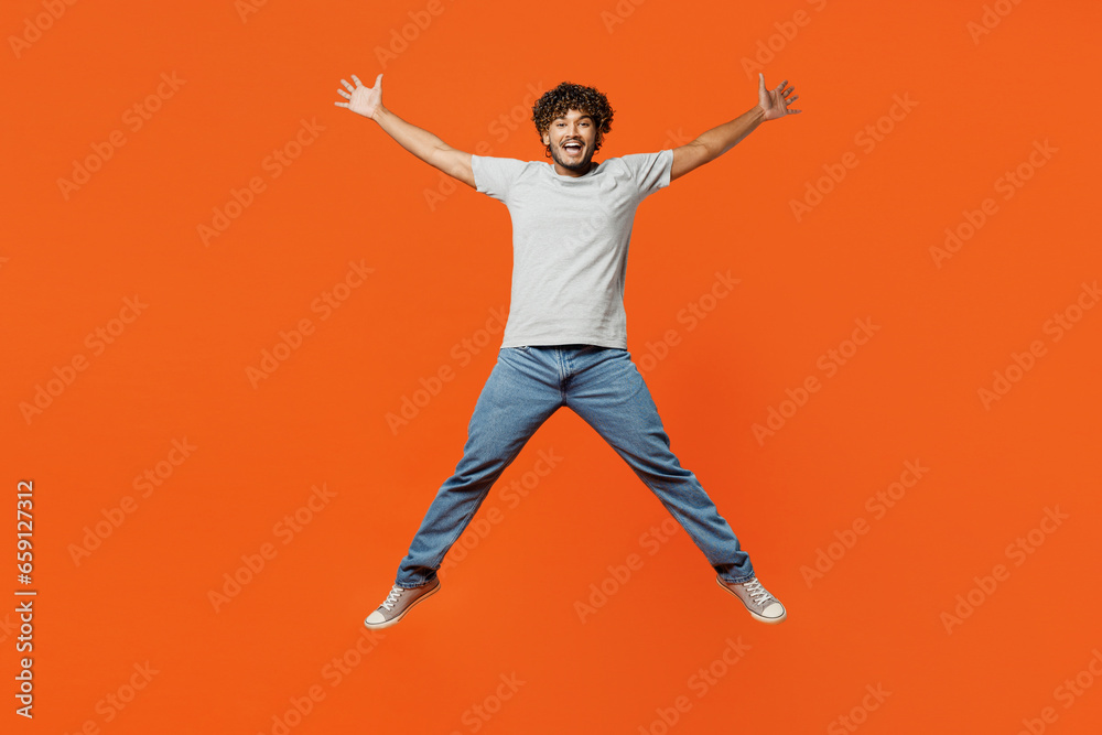 Full body young overjoyed excited smiling happy Indian man he wears t-shirt casual clothes jump high with outstretched legs hands look camera isolated on orange red color background studio portrait.