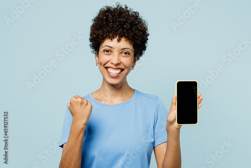 Young woman of African American ethnicity wear t-shirt casual clothes hold in hand use mobile cell phone with blank screen area do winner gesture isolated on plain pastel light blue cyan background.