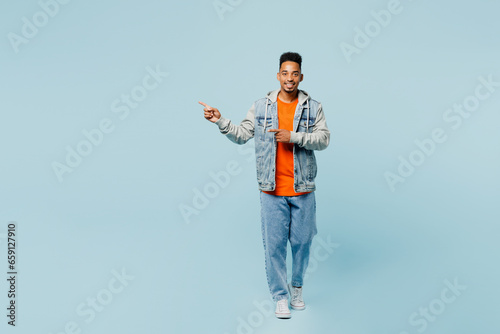 Full body fun young man of African American ethnicity wear denim jacket orange t-shirt point index finger aside indicate on workspace area mock up isolated on plain pastel light blue cyan background.