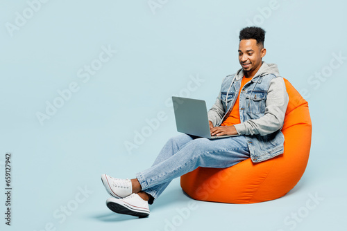 Full body young IT man of African American ethnicity wear denim jacket orange t-shirt sit in bag chair hold use work on laptop pc computer isolated on plain pastel light blue cyan background studio.