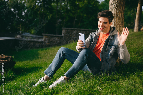 Young man wears casual clothes do selfie shot on mobile cell phone waving hand walk rest relax in spring green city park sitting on sunshine lawn outdoors on nature. Urban lifestyle leisure concept.
