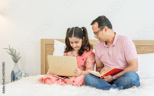 Indian happy father and daughter sitting on bed at cozy home, smiling with happiness, teaching, doing homework, playing, using laptop together. Education, Technology, Creative Activity Concept.