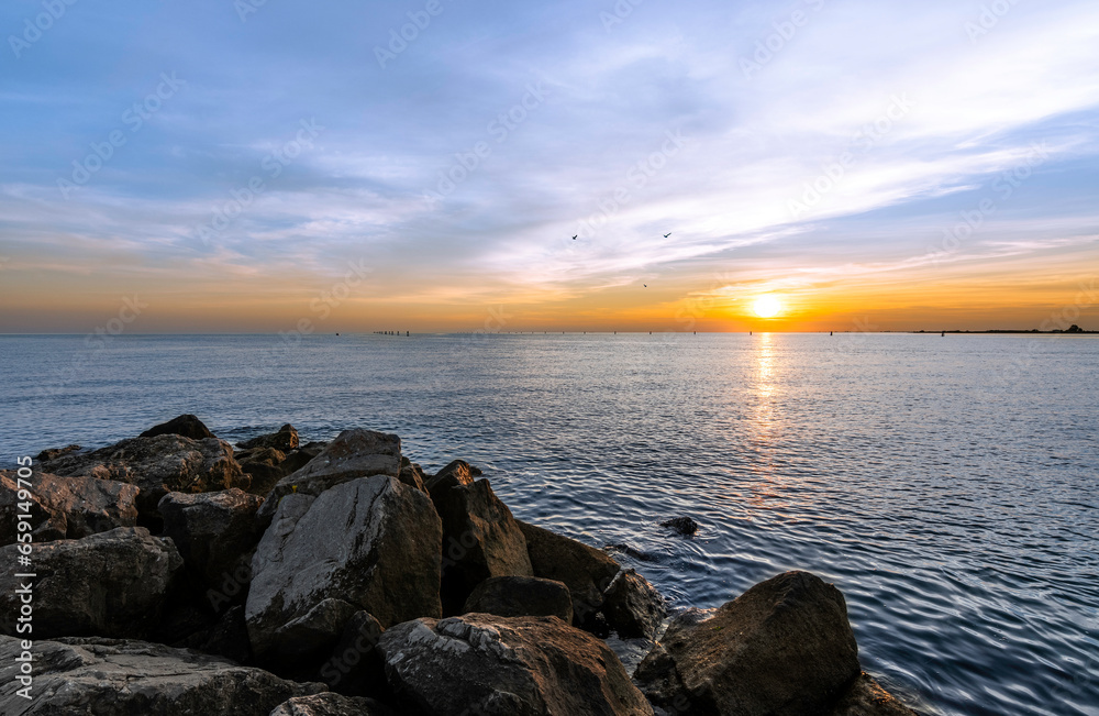 Fiery sunset taken on the rocks of a pier in Grado. The colors are warm, the sea is calm and there are no people. Three birds fly on the horizon. Multi shooting mode for HDR effect. Fiery sunset taken on the rocks of a pier in Grado. The colors are warm, the sea is calm and there are no people. Three birds fly on the horizon. Multi shooting mode for HDR effect.
