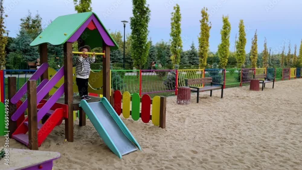 Lovely toddler in stripy jacket hanging on the horizontal bar at playground. Happy kid jumps cheerfully on top of the slide. Lovely toddler in stripy jacket hanging on the horizontal bar at playground. Happy kid jumps cheerfully on top of the slide.
