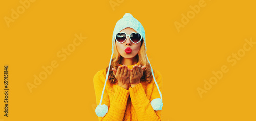 Winter portrait of beautiful woman blowing her red lips sending sweet air kiss wearing heart shaped sunglasses, yellow knitted sweater and white hat with pom pom on studio background
