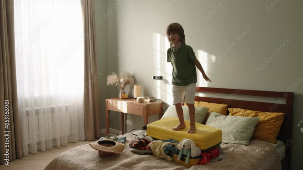 Happy kid boy jumping on suitcase. Child bouncing on packed baggage. Game fantasy, child happy to use assembled suitcase as jumping platform, trampoline, creative idea. Fun way to spend time. Happy kid boy jumping on suitcase. Child bouncing on packed baggage. Game fantasy, child happy to use assembled suitcase as jumping platform, trampoline, creative idea. Fun way to spend time.