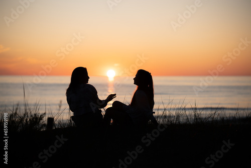 Silhouette of two female friends enjoying a conversation while sitting and watching the sunset on a beach. Back view