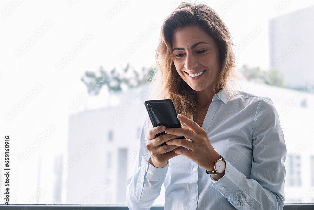 Cheerful adult female chatting on phone nearby window smiling