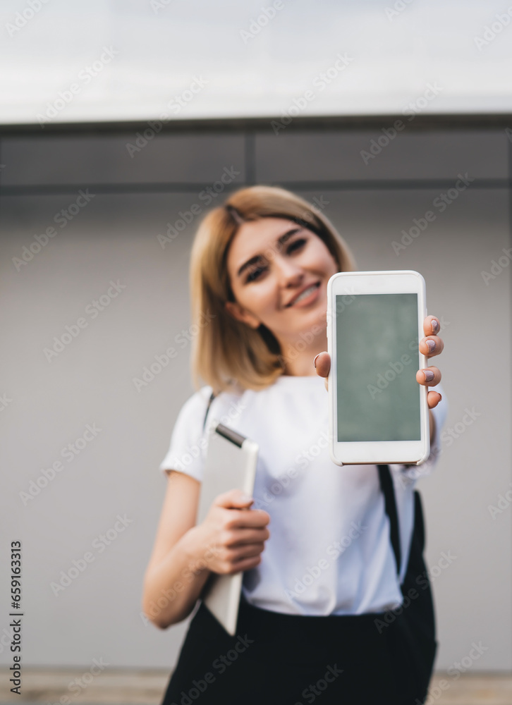 Positive female demonstrating smartphone near office building Positive female demonstrating smartphone near office building