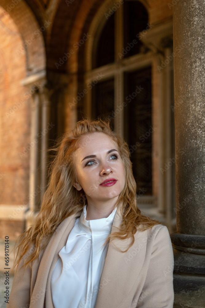 Fototapeta premium Portrait of young woman with blue eyes and golden hair in trench coat outdoors. Old city. Vertical frame.