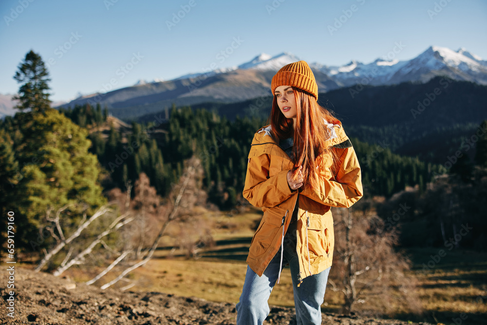 Woman hiking in the mountains in the fall with a smile with teeth and happiness in a yellow cape with red hair full-length stands against the backdrop of trees and mountains in the sunset light