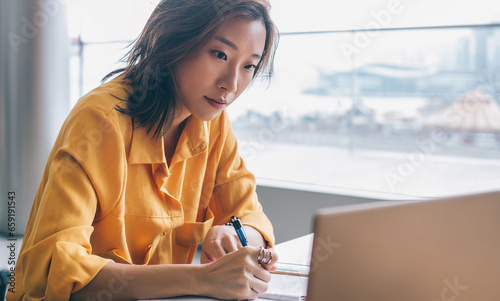 Asian female writing and looking at screen display at workplace