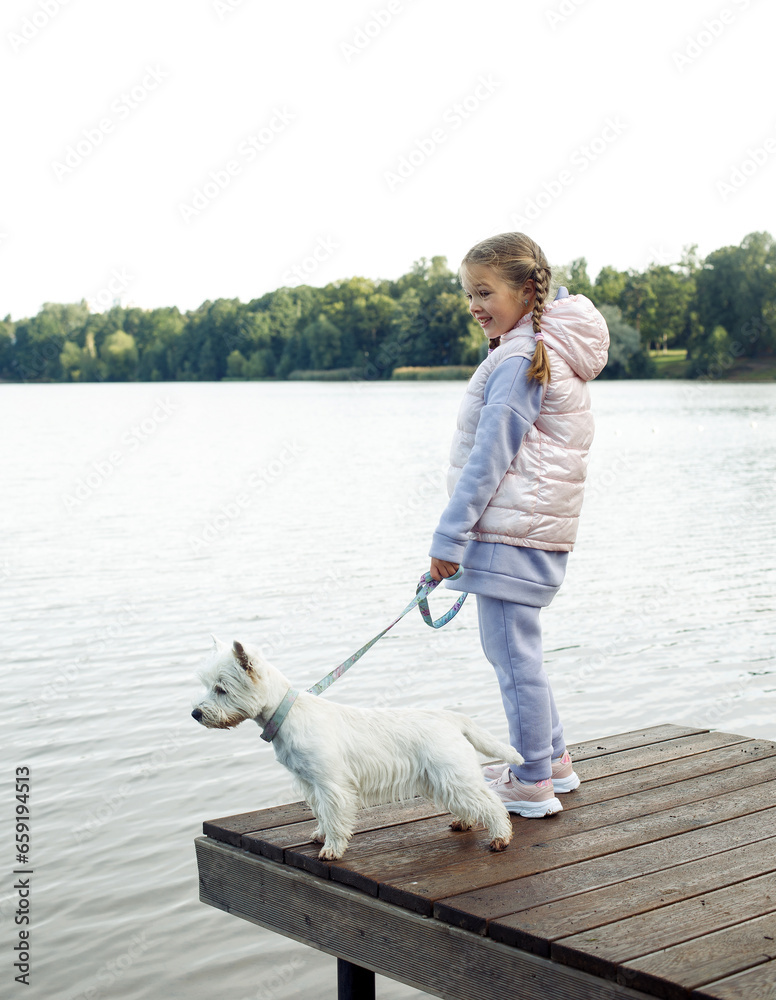 A seven-year-old girl with a small white dog stands on a bridge near the lake and looks at the water. A seven-year-old girl with a small white dog stands on a bridge near the lake and looks at the water.