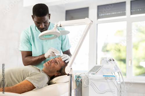 фотографія African male cosmetologist performs skin care and maintenance procedure for European man client
