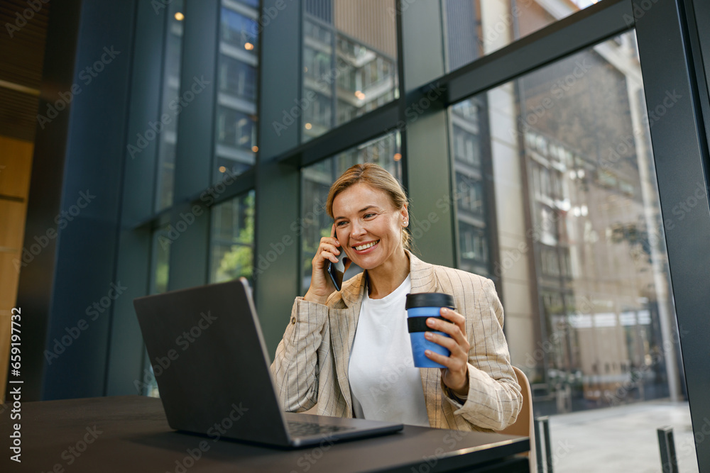 Fototapeta premium Smiling businesswoman talking on phone with client while working on laptop in office