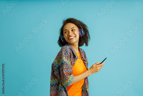 Cheerful woman in earphones with phone listen music on blue studio background and looking at side