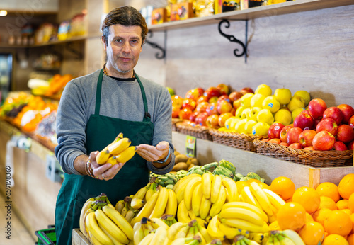 Adult man in apron sells bunches of bananas in greengrocer shop