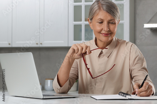 Beautiful senior woman taking notes near laptop at white marble table in kitchen
