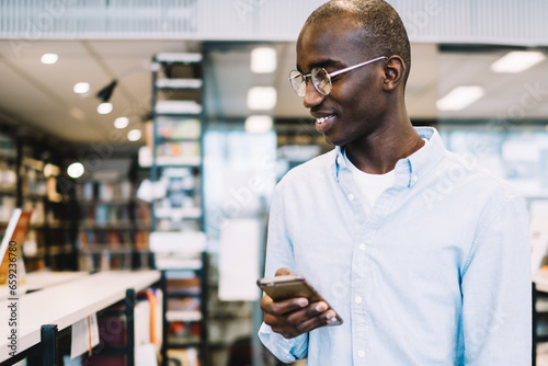 Canvas Print Black man walking along bookcases with smartphone and smiling