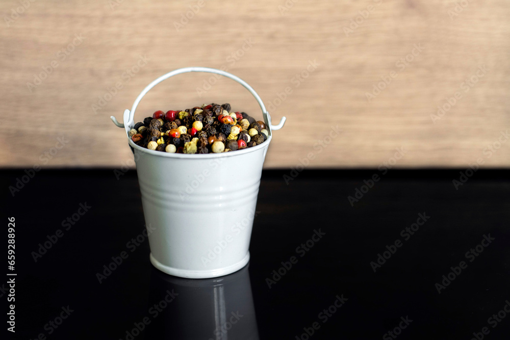 A mixture of pepper varieties with peas in the decorative little white bucket on the black background. Heap of various pepper. Mix of red, black, and white peppercorn seeds. Top view A mixture of pepper varieties with peas in the decorative little white bucket on the black background. Heap of various pepper. Mix of red, black, and white peppercorn seeds. Top view