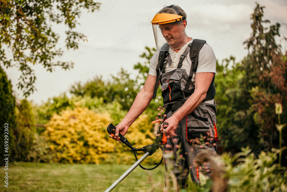An older gentleman in a safety jumpsuit demonstrating expert lawn care as he trims grass with a professional-grade trimmer in his backyard An older gentleman in a safety jumpsuit demonstrating expert lawn care as he trims grass with a professional-grade trimmer in his backyard