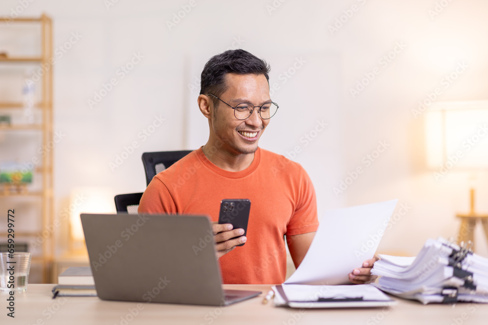 Picture of young asian man using a smartphone and smiling. working at home.accountant Documents data contract report for analysis TAX. Picture of young asian man using a smartphone and smiling. working at home.accountant Documents data contract report for analysis TAX.