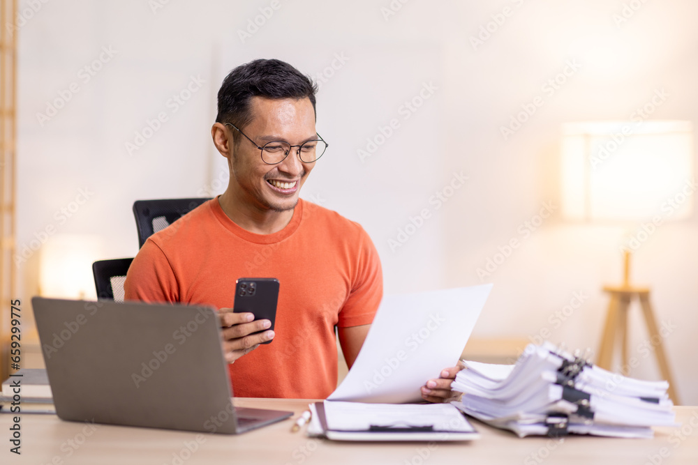 Picture of young asian man using a smartphone and smiling. working at home.accountant Documents data contract report for analysis TAX. Picture of young asian man using a smartphone and smiling. working at home.accountant Documents data contract report for analysis TAX.