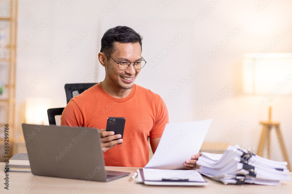 Picture of young asian man using a smartphone and smiling. working at home.accountant Documents data contract report for analysis TAX. Picture of young asian man using a smartphone and smiling. working at home.accountant Documents data contract report for analysis TAX.