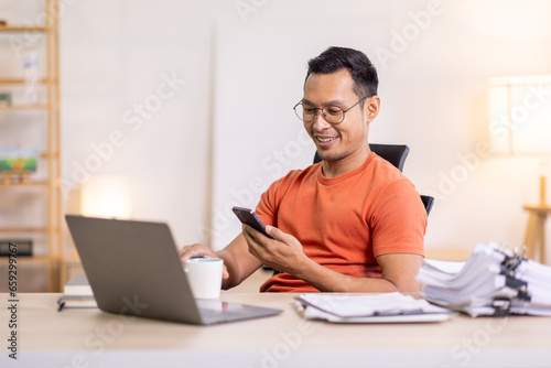 Picture of young asian man using a smartphone and smiling. working at home.accountant Documents data contract report for analysis TAX.