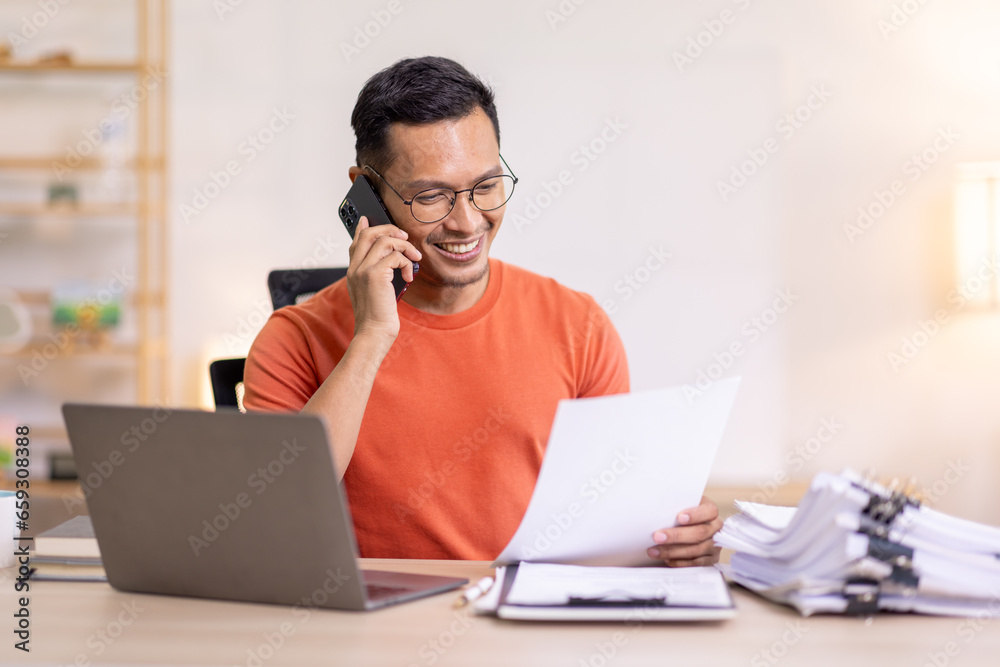 Smiling Asian man writing notes while making phone call and using laptop at home Smiling Asian man writing notes while making phone call and using laptop at home