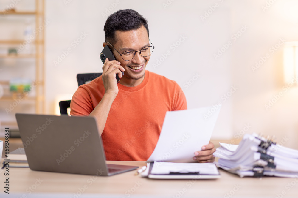 Smiling Asian man writing notes while making phone call and using laptop at home Smiling Asian man writing notes while making phone call and using laptop at home