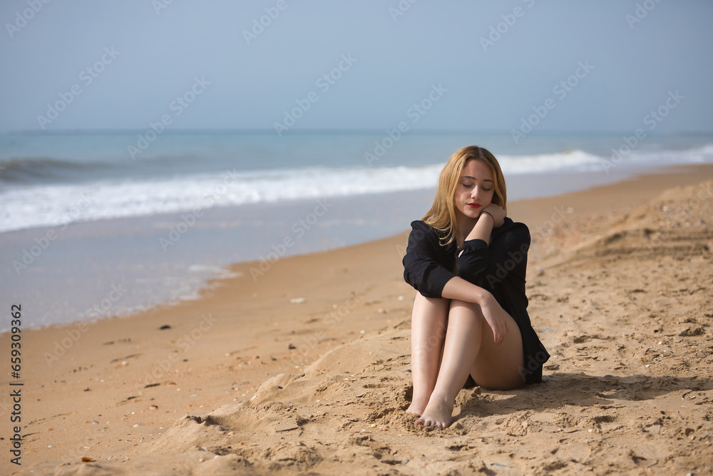 Young, blonde, beautiful woman in a bikini and black colored shirt sitting on the beach, sad, lonely, sorry, depressed. Concept loneliness, sadness, grief, depression.