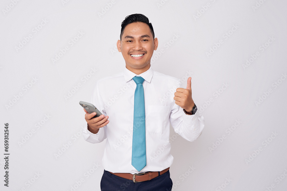 Portrait of smiling happy young Asian man in formal wear standing holding mobile phone and showing thumbs up isolated on white background
