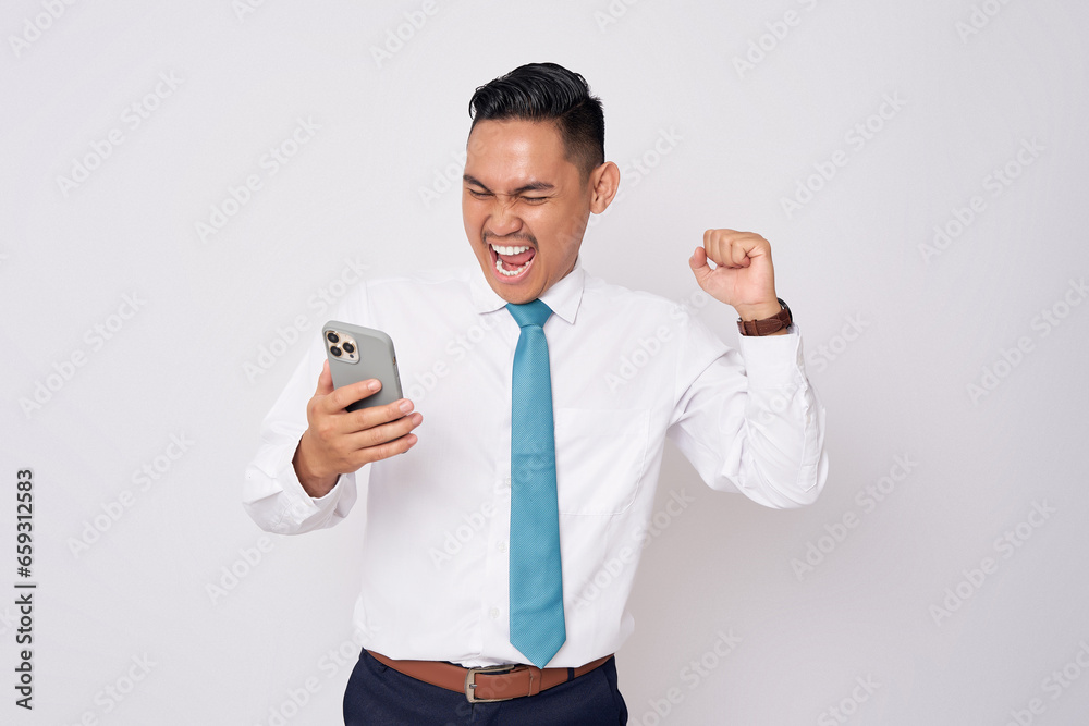 Excited  young Asian man in formal wear standing holding a mobile phone while celebrating success or winner gesture  isolated on white background