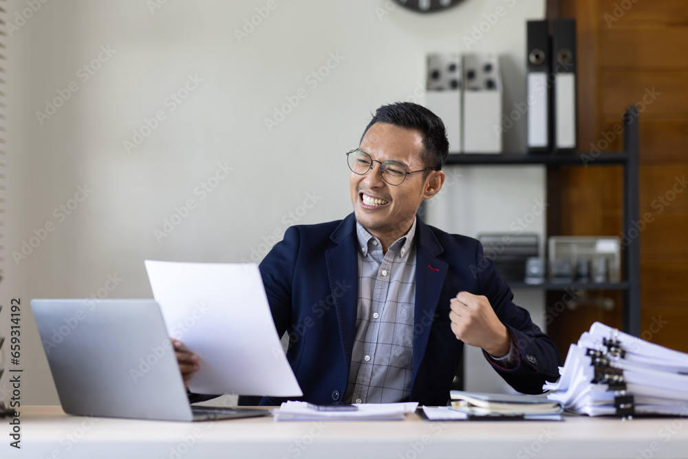 Happy Excited Young business Asian man working at workplace with laptop and papers on desk Happy Excited Young business Asian man working at workplace with laptop and papers on desk