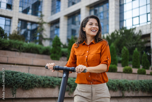 Young beautiful woman smiles and rides an electric scooter to work along office buildings. Ecological transportation concept.