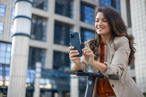 Female entrepreneur with smartphone while driving an electrical push scooter near business center on city street. Transportation environmental concept