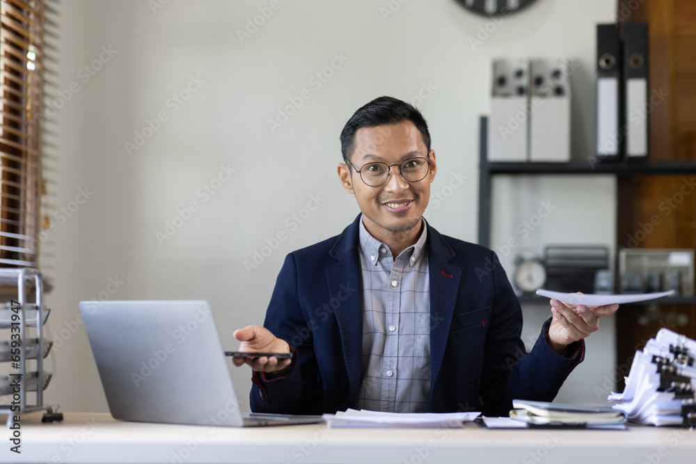 Texting to colleague or play games. Confident young asian man in smart casual wear holding smart phone and looking at it while sitting at his working place in office Texting to colleague or play games. Confident young asian man in smart casual wear holding smart phone and looking at it while sitting at his working place in office