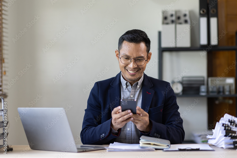 Texting to colleague or play games. Confident young asian man in smart casual wear holding smart phone and looking at it while sitting at his working place in office