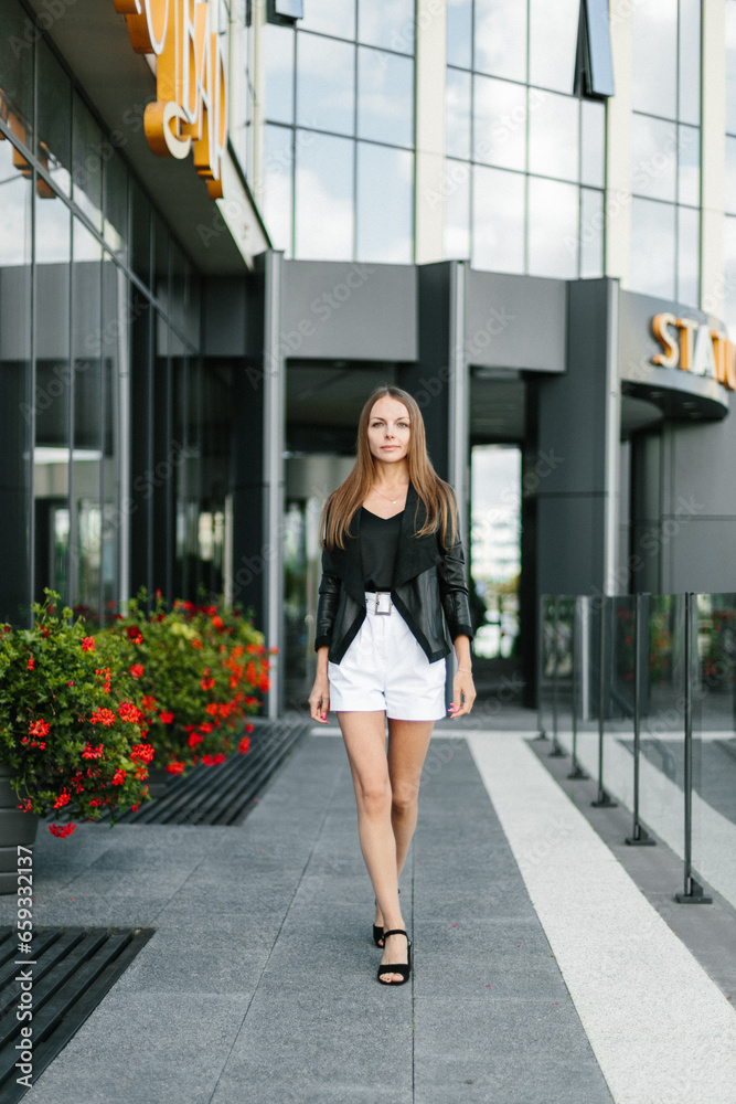 A girl with long blond hair, wearing a black top, black jacket and white shorts, poses against the backdrop of modern architecture. Young woman walking in a park with landscape design A girl with long blond hair, wearing a black top, black jacket and white shorts, poses against the backdrop of modern architecture. Young woman walking in a park with landscape design