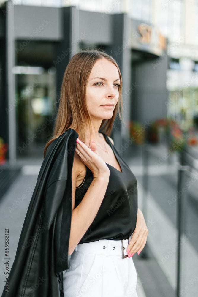 A girl with long blond hair, wearing a black top, black jacket and white shorts, poses against the backdrop of modern architecture. Young woman walking in a park with landscape design A girl with long blond hair, wearing a black top, black jacket and white shorts, poses against the backdrop of modern architecture. Young woman walking in a park with landscape design