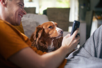 Man sitting on sofa with dog. Pet owner taking selfie through mobile phone with his Nova Scotia Duck Tolling Retriever at home..